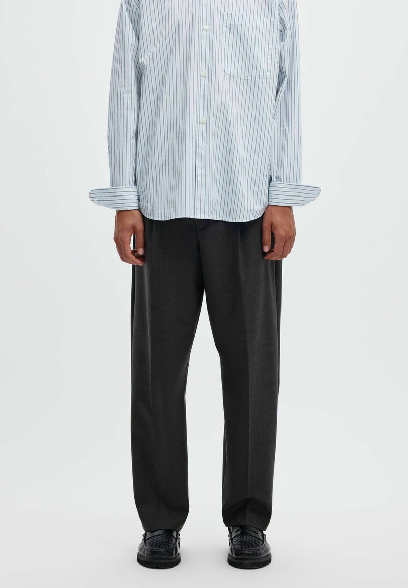 Personne portant une chemise à rayures bleu clair boutonnée, un pantalon ample gris foncé et des chaussures noires texturées, debout devant un fond uni.