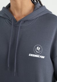 Felpa grigia realizzata in tessuto morbido con cappuccio regolabile. Il davanti presenta una grafica bianca con un volto sorridente e la scritta "DIVERTIMENTO COSMICO".