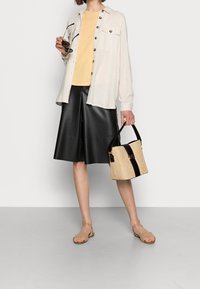 Donna che indossa una camicia beige con bottoni, una maglietta gialla, pantaloni corti in pelle nera fino al ginocchio, sandali beige, mentre tiene una borsa di paglia con tracolla nera e occhiali da sole.