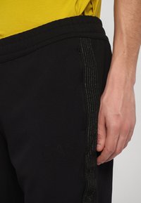 Zwarte sportieve broek met een gestructureerde afwerking, voorzien van een glanzende strook aan de zijkant en een reliëflogo op het been.