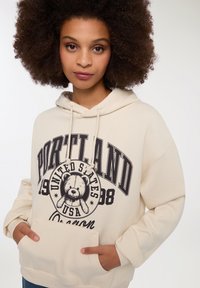 Felpa color crema con una grande stampa grafica che presenta "Portland, Oregon" e un orso. Ha una tasca a marsupio frontale e un cappuccio con cordino.