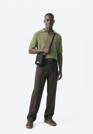 Άνδρας που φορά πλεκτό πόλο χακί, καφέ παντελόνι με ρίγες, γκρι αθλητικά παπούτσια και μαύρη τσάντα χιαστί, στέκεται μπροστά σε λευκό φόντο.