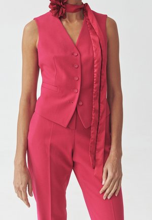 Gilet rouge sans manches sur mesure avec boutons, pantalon taille haute assorti, et une longue écharpe en satin avec une fleur en tissu nouée autour du cou.