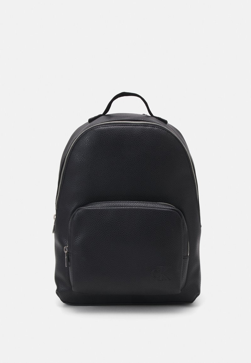 Zaino in pelle nera con superficie testurizzata, forma rotonda, tasca frontale con zip e manico superiore. Logo discreto nella parte anteriore inferiore.