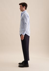 Camicia azzurro chiaro a maniche lunghe con bottoni, abbinata a pantaloni di velluto blu scuro e scarpe nere, vista di lato su uno sfondo neutro.