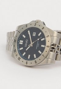 Ρολόι από ανοξείδωτο ατσάλι με σκοτεινό μπλε καντράν, ασημένιους δείκτες και παράθυρο ημερομηνίας. Περιέχει ζαρωμένη στεφάνη και μεταλλικό μπρασελέ.