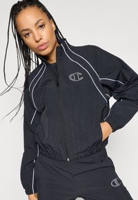 Chaqueta con cremallera negra y detalles en blanco, hecha de material ligero. Presenta puños elásticos y un logo destacado en el pecho.