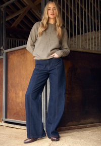 Pull en laine marron, pantalon en denim bleu foncé à jambes larges, avec poches avant et détails de couture visibles, associé à des bottes en cuir marron.