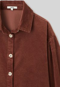 Chemise en velours côtelé marron avec un col, cinq boutons sur le devant et un tissu texturé avec des motifs côtelés verticaux.