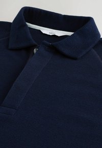 Navy blue polo shirt made of textured fabric with a buttoned placket, short collar, and a branded tag labeled "NEXT."