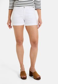 Shorts de mezclilla blanca con dobladillos enrollados, con cierre delantero de botón y dos bolsillos frontales, combinados con mulas de tacón abierto marrones.