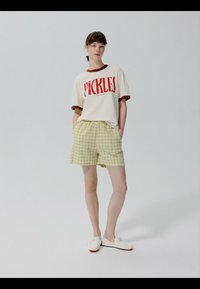 Mladá žena stojí s rukami v kapsách, má na sebe biele tričko s nápisom "PICKLES" červenou farbou, žlté kárované šortky a biele topánky na sivom pozadí.