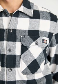 Gros plan sur une chemise à carreaux noir et blanc avec une poche à rabat boutonée arborant un petit patch logo Dickies.