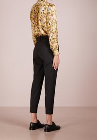 Žlutá saténová blůza s květinovým vzorem a dlouhými rukávy, v kombinaci s černými zúženými kalhotami a černými botami, pohled zezadu.