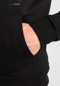 Schwarzes Hoodie mit gerippten Bündchen und Tasche. Weiche Stofftextur. Auffällige Nahtdetails am Rand der Tasche.
