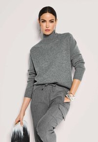 Eine Frau mit dunklen Haaren trägt einen grauen Rollkragenpullover, graue Cargo-Hosen, silberne Ohrringe und Armbänder und hält eine schwarz-weiße Handtasche.