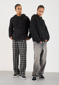 Twee modellen in zwarte gebreide hoodies. Eén met zwart-witte geblokte broek, de ander in grijze spijkerbroek. Beiden dragen zwarte sneakers.