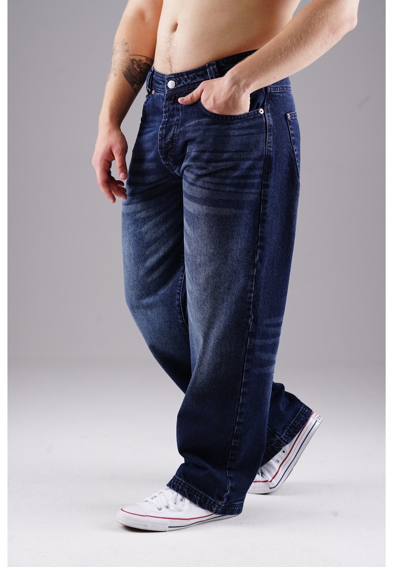 Dunkelblaue Jeans aus Denim mit entspannter Passform, geradem Bein und sichtbaren Nähten. Mit zwei Seitentaschen. Getragen mit weißen Sneakers.