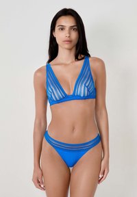 Niebieski zestaw bielizny składający się z przezroczystego, wzorzystego biustonosza o głębokim dekolcie w kształcie V oraz pasujących majtek niskich. Oba elementy mają dekoracyjne akcenty.