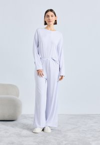 Svetlo vijoličen komplet za sproščanje, ki vključuje dolgo majico z okroglim izrezom in gumbi ter široke hlače.