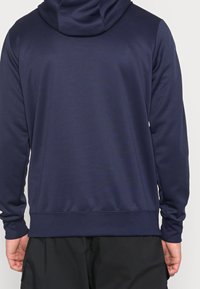 Mies, joka pukeutunut yksiväriseen tummansiniseen huppariin ja mustiin shortseihin, kuvattu takaapäin, seisoo yksinkertaisen vaalean taustan edessä.