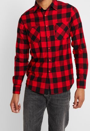 Man wearing red and black checkered long-sleeve button-up shirt with two chest pockets and dark gray jeans, cropped at the neck.