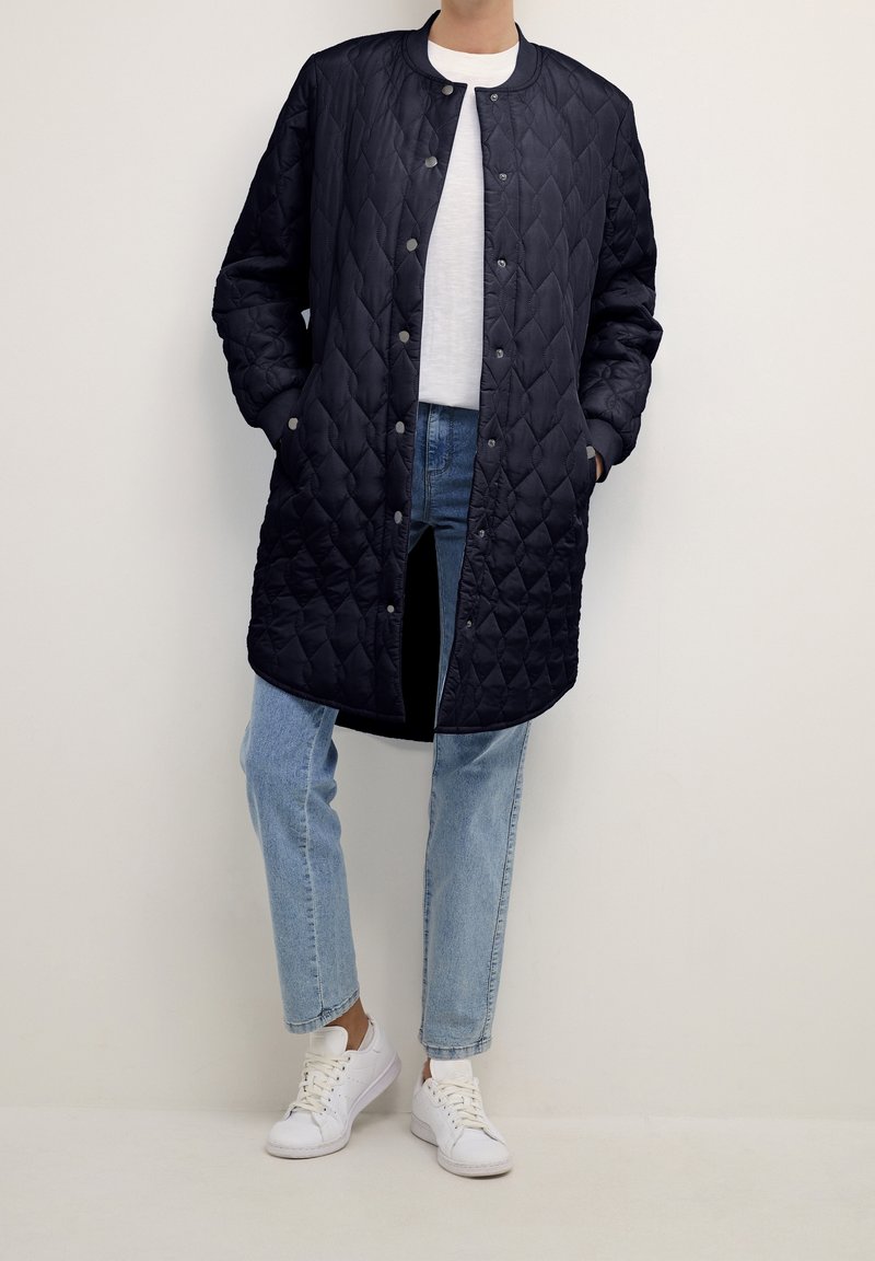 Personne portant un long manteau matelassé noir sur une chemise blanche, un jean bleu clair et des baskets blanches, debout contre un mur uni.