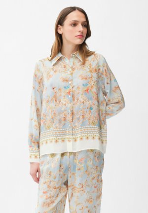 Frau trägt hellblaues Hemd mit Knöpfen und passende Hose mit intricate Orangen- und Goldblumen-Paisley-Mustern, steht vor einfachem Hintergrund.