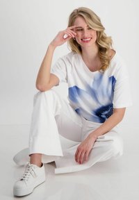 Weißes T-Shirt mit blauem Blumenmuster; lockerer Schnitt. Kombiniert mit weißen, weiten Hosen und weißen Plateau-Sneakern. Glatte Textur.