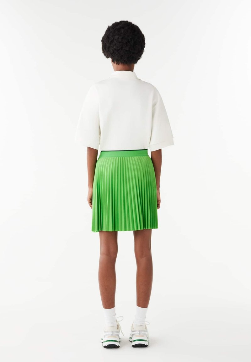Biała bluzka z krótkim rękawem i zielona plisowana spódnica, z dopasowanym pasem i fakturowanym materiałem. Sneakersy i skarpetki dopełniają stylizację.