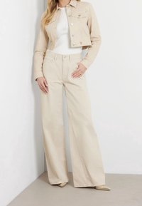 Beige Crop-Jeansjacke mit zwei Fronttaschen, kombiniert mit weiten beigefarbenen Hosen. Das Model trägt ein enges weißes Oberteil und beigefarbene Schuhe.