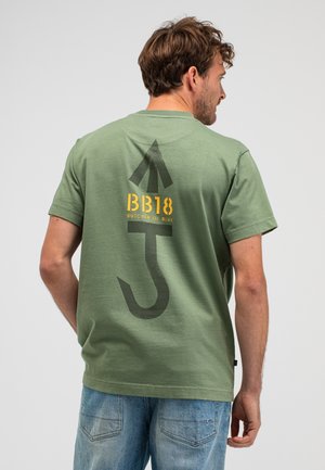 Man draagt een groen T-shirt met een grote zwarte pijl en de tekst "BB18 Butcher of Blue" op de achterkant, gecombineerd met lichtblauwe jeans.
