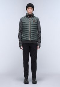 Grüne gesteppte Weste mit hohem Kragen, graues Langarmshirt darunter, schwarze Hose und Strickmütze. Gepolsterte Textur mit seitlichen Akzenten.