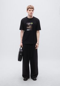 Μαύρο μπλουζάκι με γραφικό σχέδιο, συνδυασμένο με φαρδιά μαύρη παντελόνια και μαύρες παντόφλες. Το μοντέλο κρατάει μια μαύρη τσάντα χειρός με κομψό σχεδιασμό.