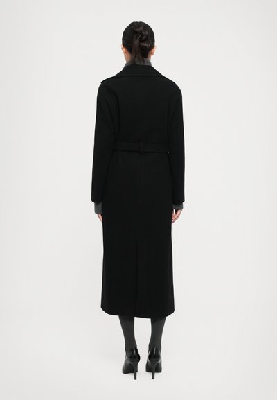 Woman with dark hair in a bun wearing a long black belted coat, gray sweater, gray leggings, and black high-heeled shoes, facing away.