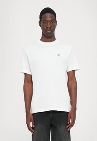 Hvit bomull T-skjorte med korte ermer, rund hals og liten brodert logo på venstre bryst; kombinert med mørkejeans, minimalistisk design.