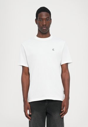 Hvit bomull T-skjorte med korte ermer, rund hals og liten brodert logo på venstre bryst; kombinert med mørkejeans, minimalistisk design.