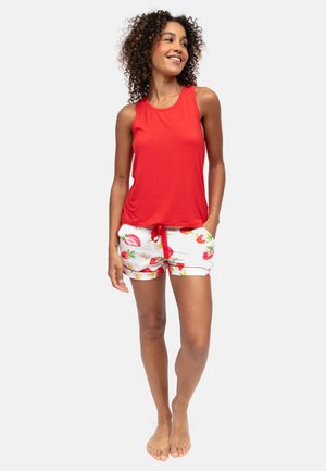 Donna in piedi a piedi nudi, indossa un top rosso senza maniche e pantaloncini bianchi con stampe di fragole rosse, sorride e guarda di lato.