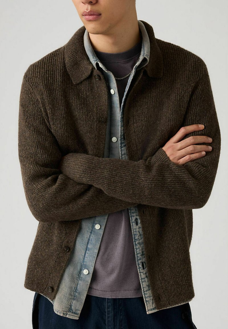 Cardigan en tricot côtelé marron avec boutons, design à col, porté par-dessus une chemise en denim bleu clair, avec une coupe décontractée et un tissu texturé.