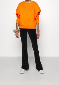 Felpa arancione con colletto a girocollo e logo ricamato, abbinata a pantaloni neri svasati e sneakers bianche. Scrunchie al polso.