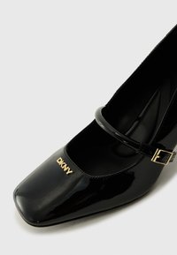 Black patent leather shoe with square toe, ankle strap, and gold "DKNY" logo on the front. Smooth texture and sleek design.