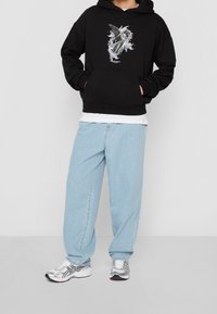 Černá mikina s kapucí s grafickou potiskem, v kombinaci s volnými světle modrými džínami a stříbrnými běžeckými botami. Styl ležérního městského oblečení.