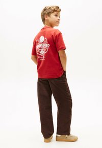 Rød bomulds t-shirt med hvidt trykt tekst og grafik på ryggen, parret med løse brune fløjlsbukser og beige sneakers.