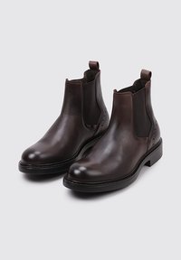 Bottes Chelsea en cuir marron avec des panneaux élastiques sur les côtés, des orteils arrondis et des tirettes. Texture lisse et finition légèrement brillante.