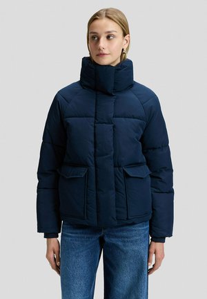 Frau trägt eine dunkelblaue Steppjacke mit großem Kragen, Klappentaschen und blauen Jeans, steht vor einem schlichten, hellgrauen Hintergrund.