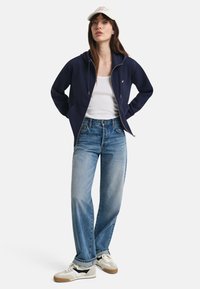 Mørkeblå hettegenser med glidelås, hvit tanktop, lyseblå jeans og beige sneakers. Jakken har hette og frontlommer. Capsen er hvit.