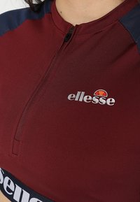 Burgundin urheilupaita, jossa on vetoketjukaulus, musta joustokappale, jossa on "ellesse"-teksti, sekä tummansinisiä hartia-akselit. Sujuva, joustava kangas.