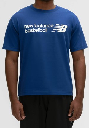 Homme portant un t-shirt bleu à manches courtes avec texte et logo blancs "new balance basketball", associé à un pantalon noir.