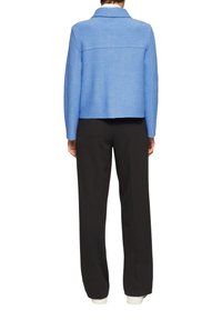 Jasnoniebieska kurtka z wełny w połączeniu z innymi materiałami, z kołnierzem, prostym krojem i przeszyciami na plecach, zestawiona z czarnymi spodniami i białymi mankietami.