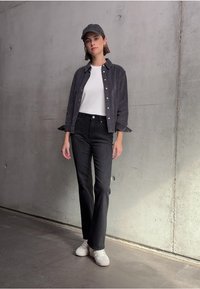 Grijze corduroy overhemd, witte t-shirt, zwarte spijkerbroek, en witte sneakers. Het overhemd heeft knopen en opgerolde mouwen, tegen een betonnen achtergrond.